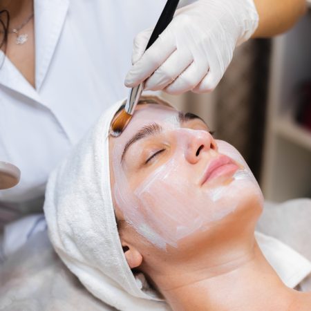 Beautician with a brush applies a white moisturizing mask to the face of a young girl client in a spa beauty salon
